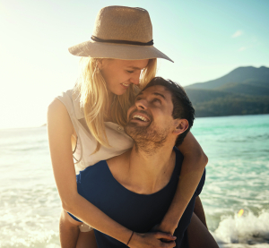 Lachendes Paar am Strand &ndash; sie tr&auml;gt einen Strohhut, er tr&auml;gt sie huckepack, im Hintergrund Meer und Berge.