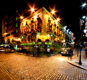 Ein traditioneller irischer Pub in Dublin bei Nacht, hell erleuchtet mit bunten Fassaden und Fahnen, Menschen flanieren über das Kopfsteinpflaster.