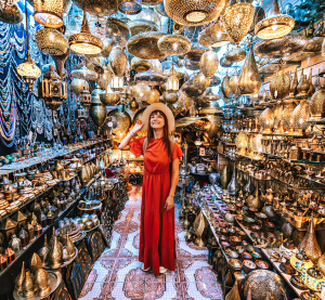 Frau in rotem Kleid steht in einem orientalischen Lampenladen voller goldener Leuchten und Kunsthandwerk.