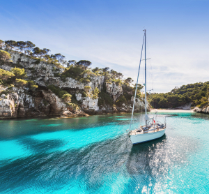 Segelboot in einer t&uuml;rkisblauen Bucht auf Menorca, umgeben von steilen Felsklippen mit Pinienbewuchs und klarem Himmel dar&uuml;ber.