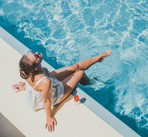 Frau sitzt entspannt am Rand eines Pools und h&auml;lt die F&uuml;&szlig;e ins Wasser, ein Getr&auml;nk steht neben ihr.
