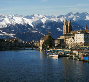 Z&uuml;richer Altstadt an der Limmat mit den Kircht&uuml;rmen und schneebedeckten Alpen im Hintergrund.