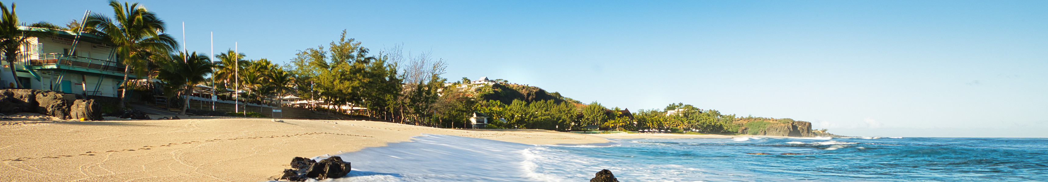 Wellen rollen &uuml;ber den goldenen Sandstrand von Saint-Gilles-les-Bains auf der Insel La R&eacute;union, umgeben von Palmen und K&uuml;stenlandschaft unter klarem, blauem Himmel.