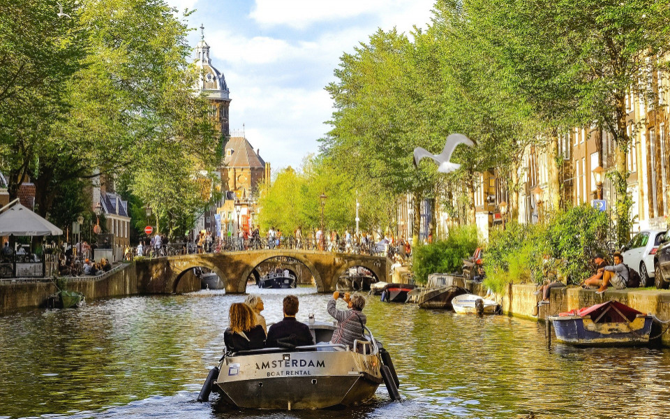 Boot mit Touristen auf einer Grachtenrundfahrt durch das grüne Kanalnetz von Amsterdam mit Blick auf eine historische Brücke und Kirchturm.