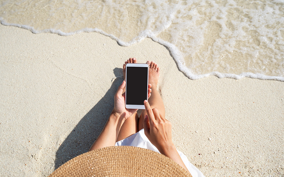 Frau sitzt mit einem Smartphone in der Hand am Sandstrand direkt am Wasser, ihre Füße berühren den nassen Sand