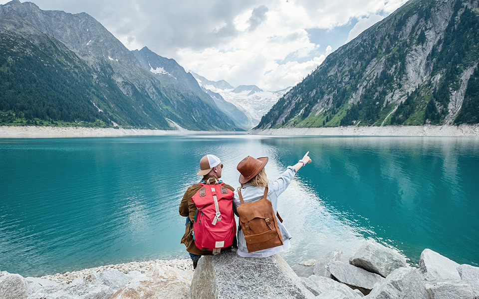 Zwei Wanderer mit Rucksäcken sitzen am Ufer eines türkisfarbenen Bergsees inmitten der Alpen.