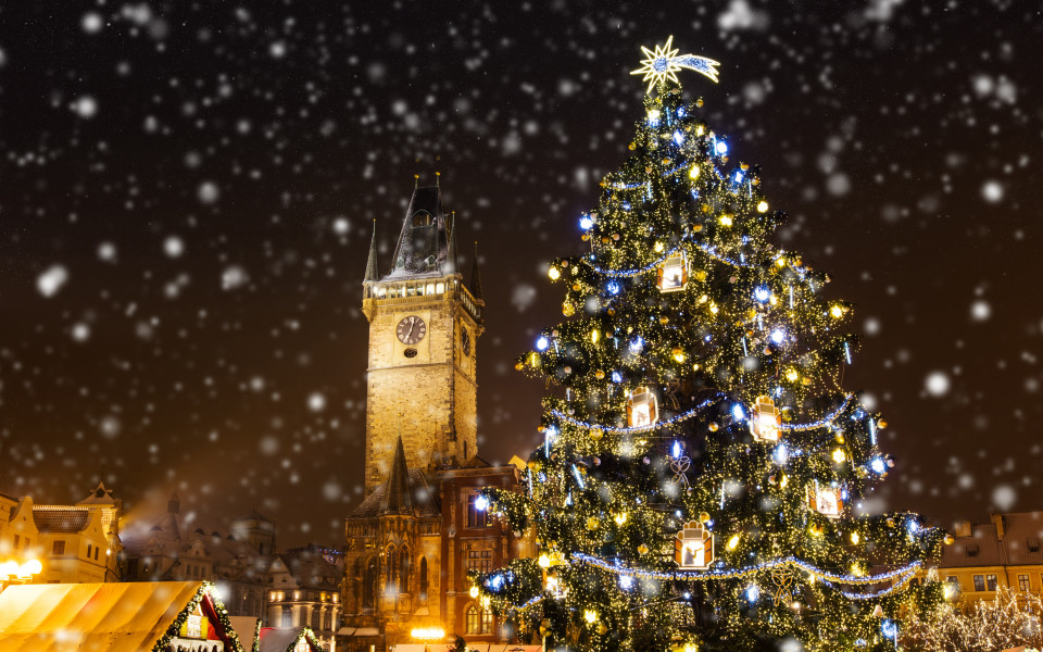 Festlich beleuchteter Weihnachtsbaum auf dem Weihnachtsmarkt in Prag, einem der sch&ouml;nsten Adventsm&auml;rkte weltweit, mit dem historischen Uhrturm im Hintergrund und sanft fallendem Schnee.
