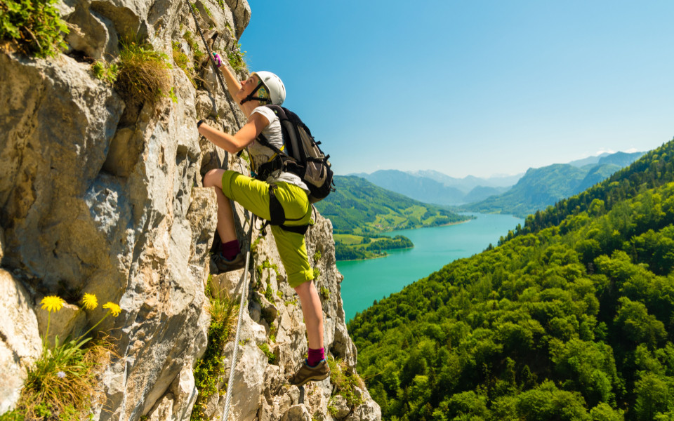 Kletterin sichert sich am Felsen mit Klettersteigset, im Hintergrund liegt der t&uuml;rkisfarbene Attersee im Salzkammergut.