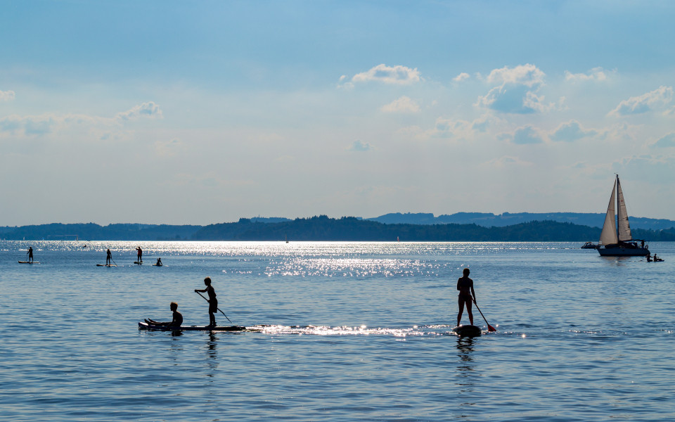 Wassersport am Chiemsee mit Personen, die auf Stand-Up-Paddle-Boards fahren, w&auml;hrend ein Segelboot im Hintergrund f&auml;hrt.