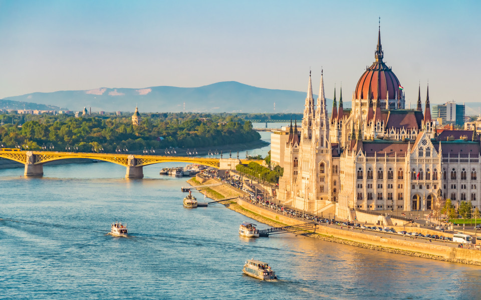 Ungarisches Parlamentsgeb&auml;ude in Budapest, an der Donau mit Booten und einer Br&uuml;cke im Hintergrund.