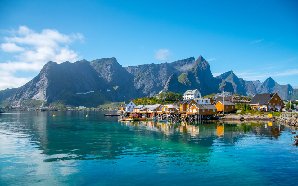 Ein Fischerdorf in der Nähe von Reine auf den Lofoten mit traditionellen Holzhäusern am Wasser, umgeben von einer Berglandschaft.