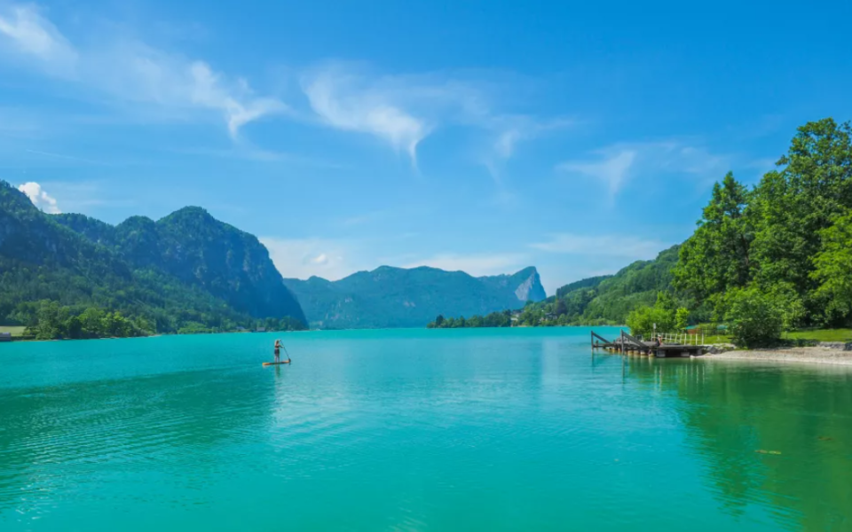 Stand-up-Paddler auf t&uuml;rkisblauem See im Salzkammergut, umgeben von bewaldeten Bergh&auml;ngen und Steg am rechten Ufer.