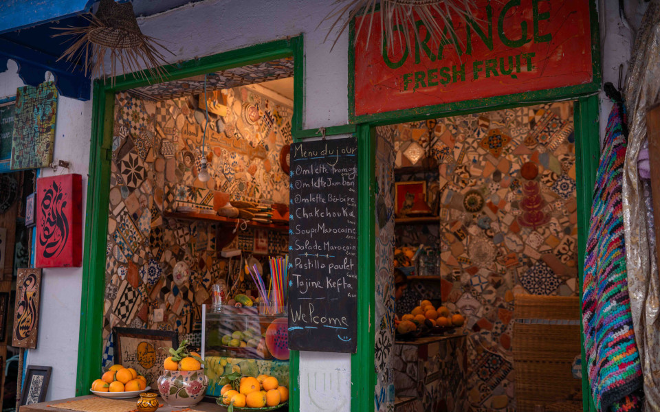 Kleines buntes Lokal in Essaouira mit handbemalter Mosaik-Dekoration, frischen Orangen und Tafel mit marokkanischen Gerichten wie Tajine und Pastilla.