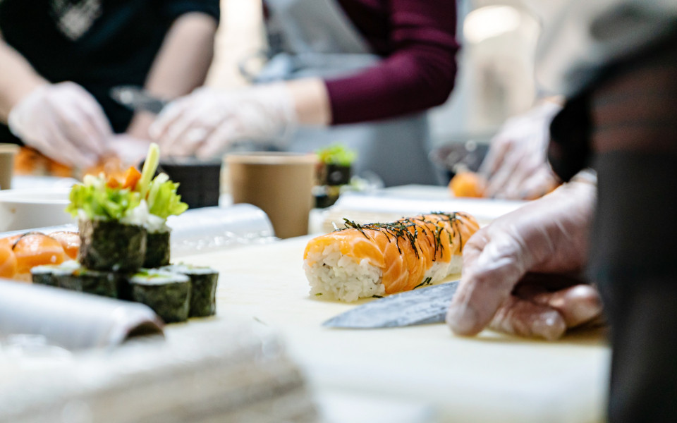 Mehrere Personen bereiten an einer Theke Sushi zu; im Vordergrund liegt eine frisch gerollte Lachsrolle mit Reis und Nori, daneben weitere Sushi-Stücke und Zutaten auf einem hellen Schneidebrett.